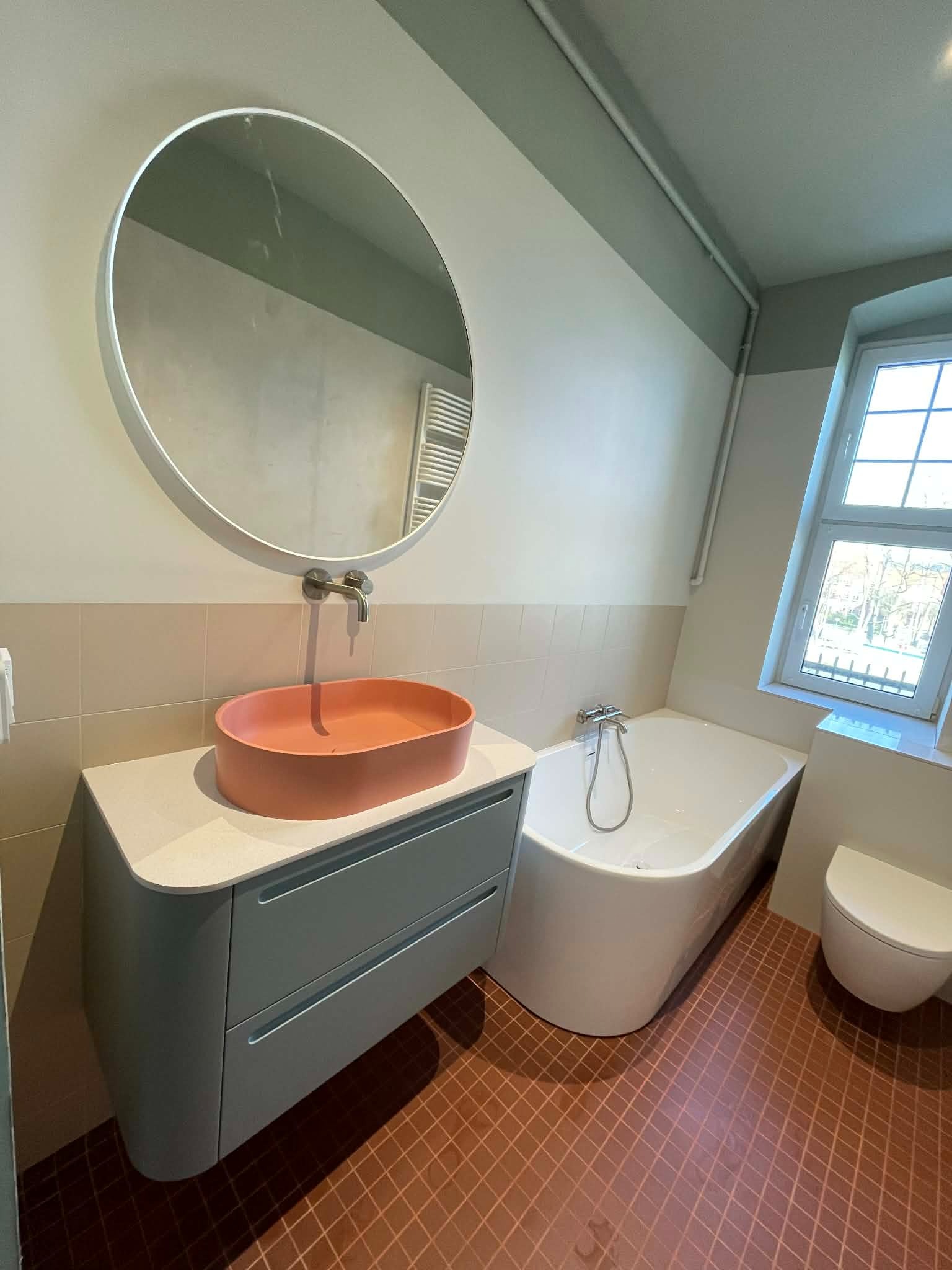 Nowoczesna łazienka z okrągłym lustrem, pomarańczową umywalką na szafce w kolorze gołębim, białą wanną i terakotową podłogą w kratkę. Jasne, naturalne światło.