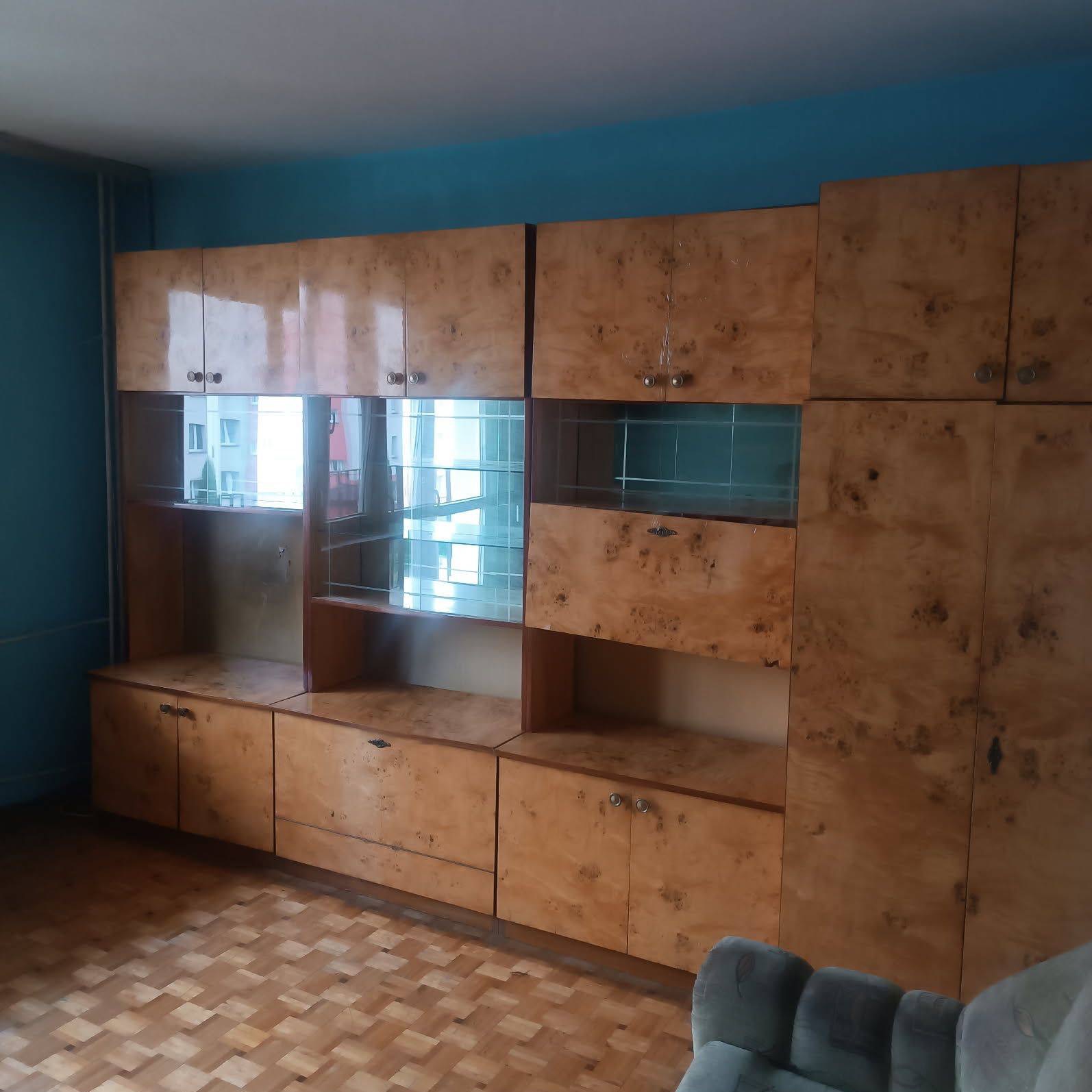 Masywna, fornirowana meblościanka w stylu retro z przeszklonymi witrynami, na tle turkusowej ściany i parkietu. Widoczny fragment szarej sofy.