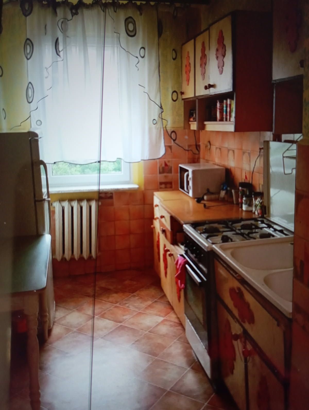 Kuchnia z brązowymi płytkami i wzorzystymi szafkami w Pionkach. Widok na okno z firanką i grzejnik. Sprzęty kuchenne na blacie.