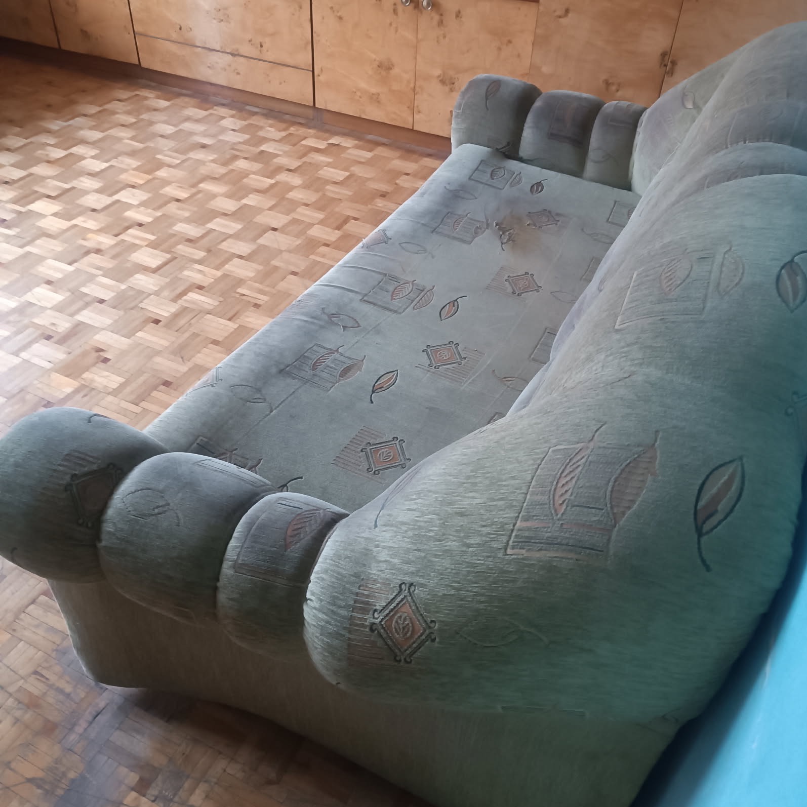 Używana, zielonkawa sofa z wzorem liści i rombów na tle parkietu i drewnianej szafki. Widoczne plamy na tapicerce.