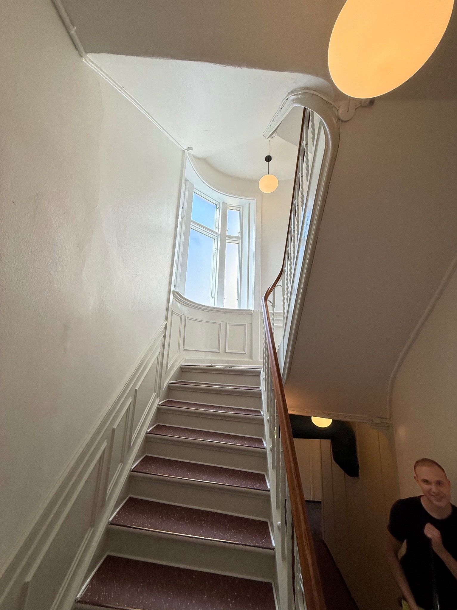 Klatka schodowa z białymi ścianami i schodami z bordową wykładziną. Na końcu schodów okno z widokiem na niebo. W rogu widoczna osoba.