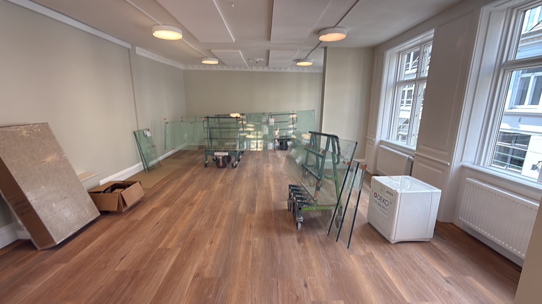Puste pomieszczenie z drewnianą podłogą, przygotowane do remontu. Widoczne zabezpieczone szkła na wózkach, kartony i biały pakunek. Duże okna w tle.