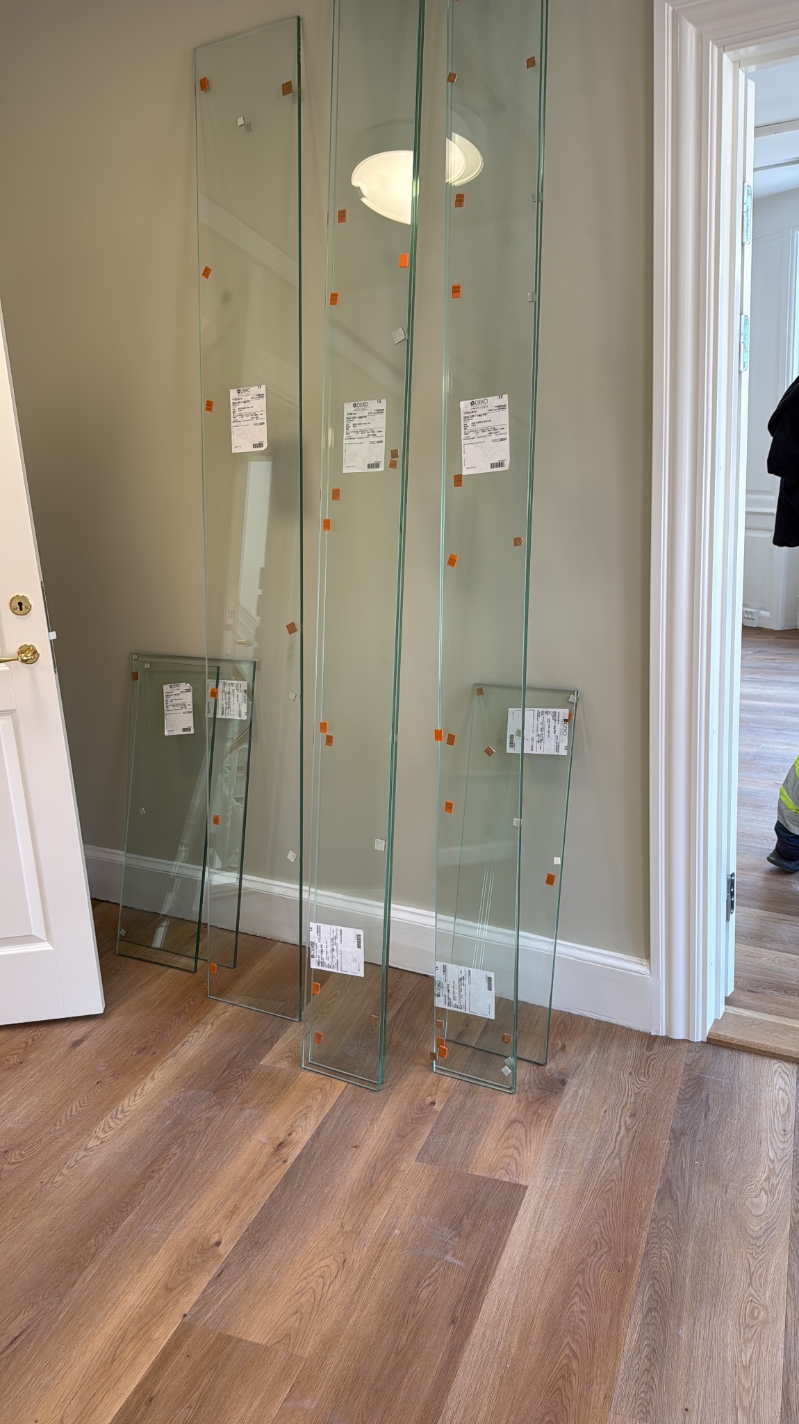 Szyby szklane z etykietami, ustawione pionowo na drewnianej podłodze w jasnym wnętrzu. Widoczne fragmenty drzwi i osoby w tle.