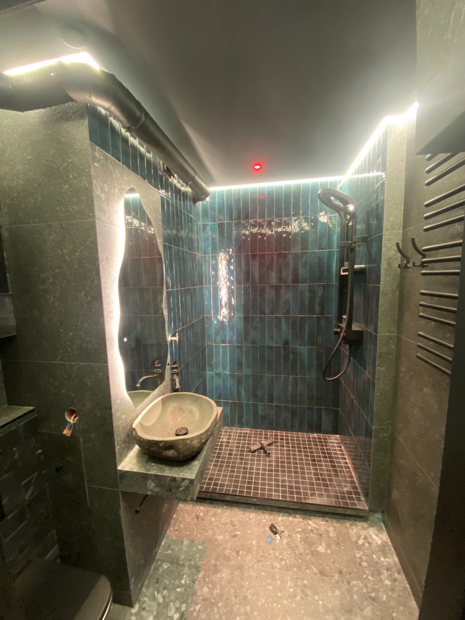 Nowoczesna łazienka z turkusowymi płytkami, kamienną umywalką i podświetlanym lustrem. Ciemna kolorystyka, minimalistyczny design, widoczne elementy instalacji.