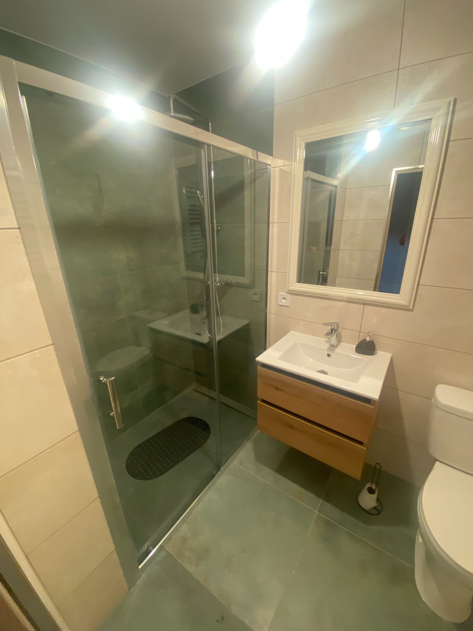 Nowoczesna łazienka z kabiną prysznicową ze szklanymi drzwiami, umywalką z drewnianą szafką i dużym lustrem w białej ramie. Beżowe płytki na ścianach i szare na podłodze.