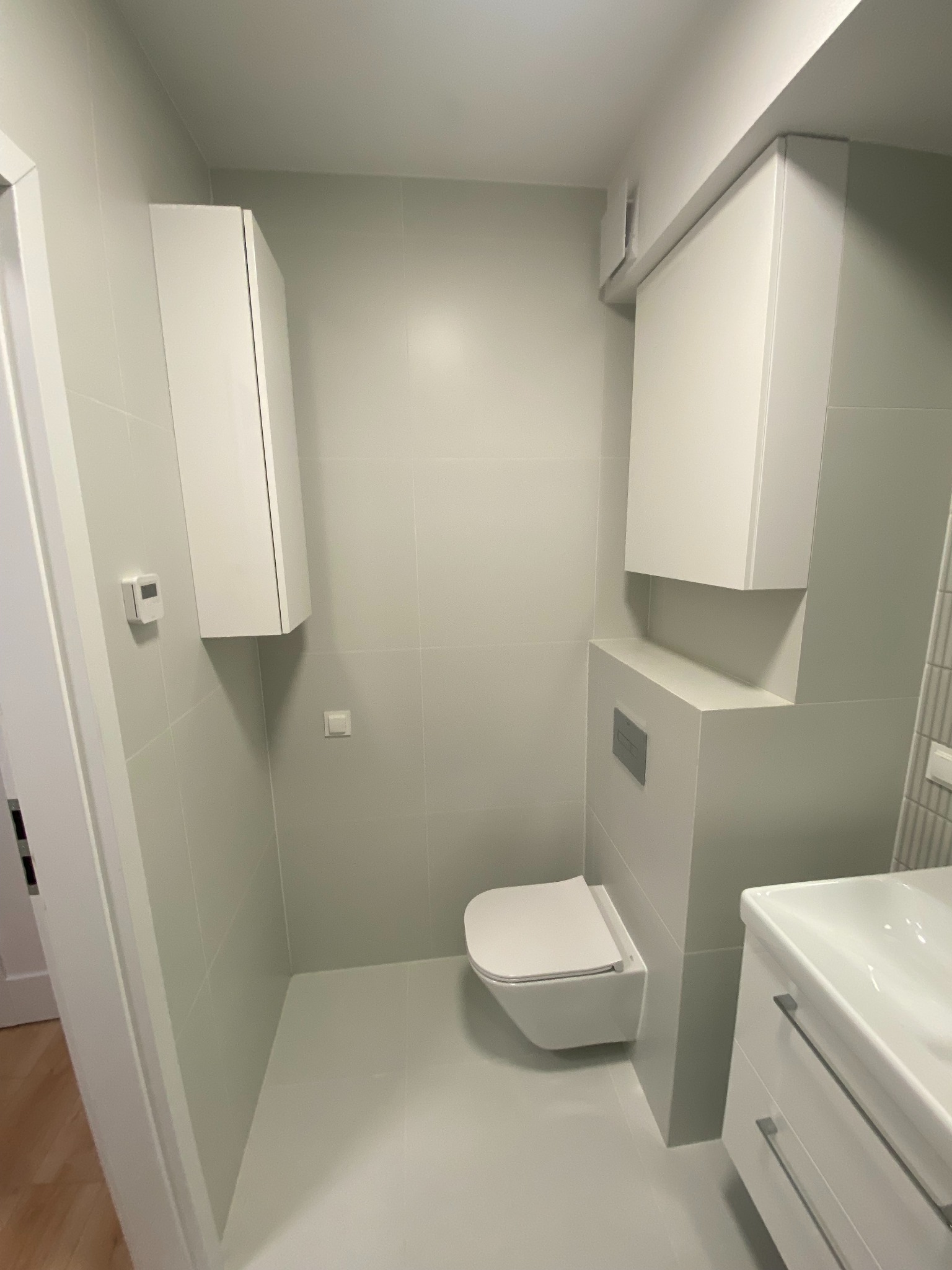 Nowoczesna, minimalistyczna łazienka w odcieniach szarości z podwieszaną toaletą, szafkami i umywalką. Widoczne płytki ceramiczne i armatura.