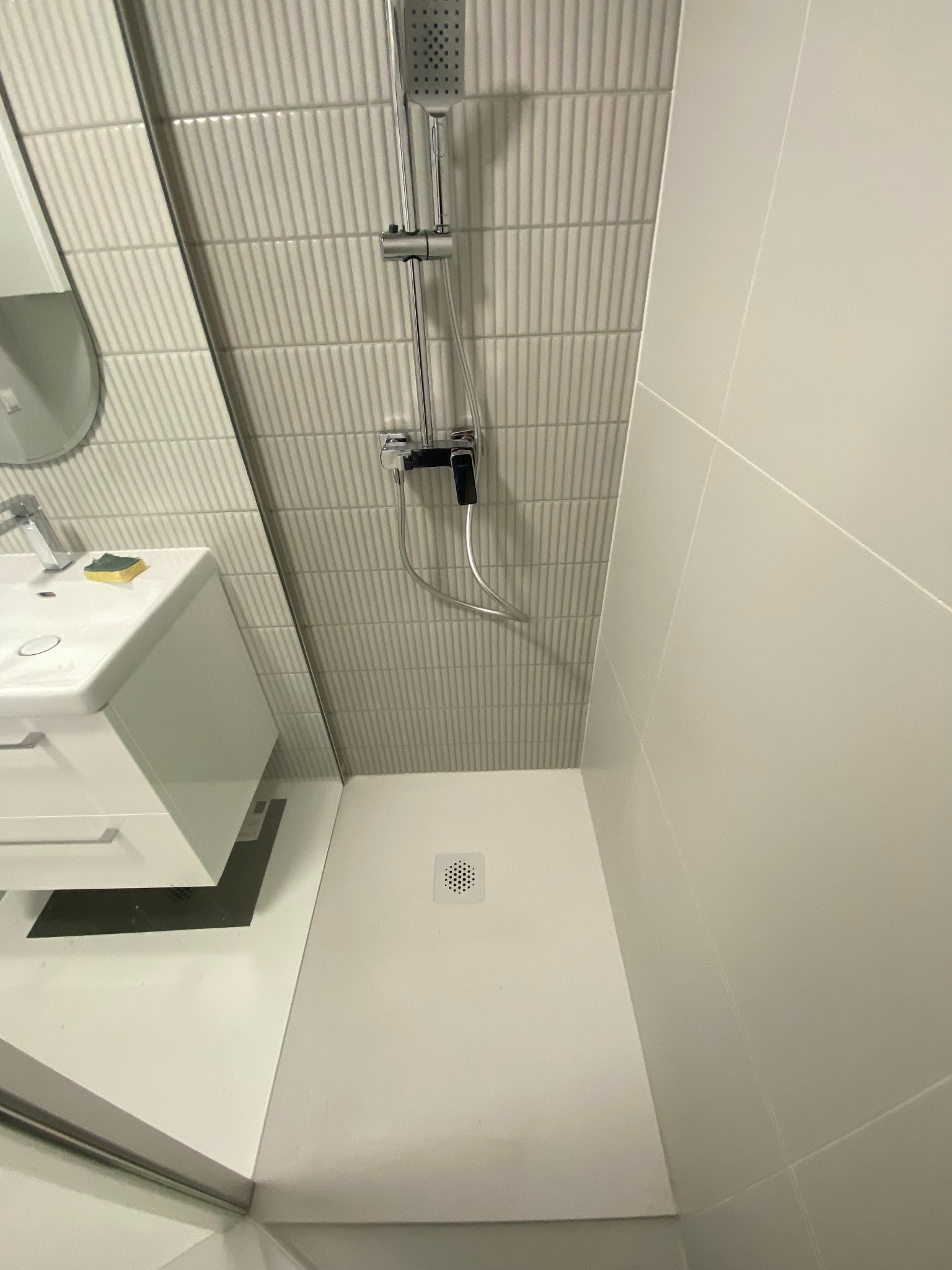 Nowoczesna, minimalistyczna łazienka po remoncie: białe płytki, kabina prysznicowa typu walk-in, umywalka z szafką. Widoczny odpływ liniowy i bateria prysznicowa.