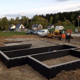 Roboty ziemne usługi koparką - Zabezpieczony fundament domu jednorodzinnego z widoczną hydroizolacją, na tle krajobrazu z domami i sprzętem budowlanym. Dwóch mężczyzn stoi obok pomarańczowej mini koparki.
