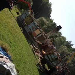 Roboty ziemne usługi koparką - Demolka budynku mieszkalnego przez koparkę na zielonym terenie. Widoczne fragmenty konstrukcji i maszyny budowlanej. Prace rozbiórkowe w toku.