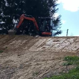 Roboty ziemne usługi koparką - Pomarańczowa mini koparka na nierównym, piaszczystym terenie z zieloną trawą w tle, wykonująca prace ziemne w słoneczny dzień.