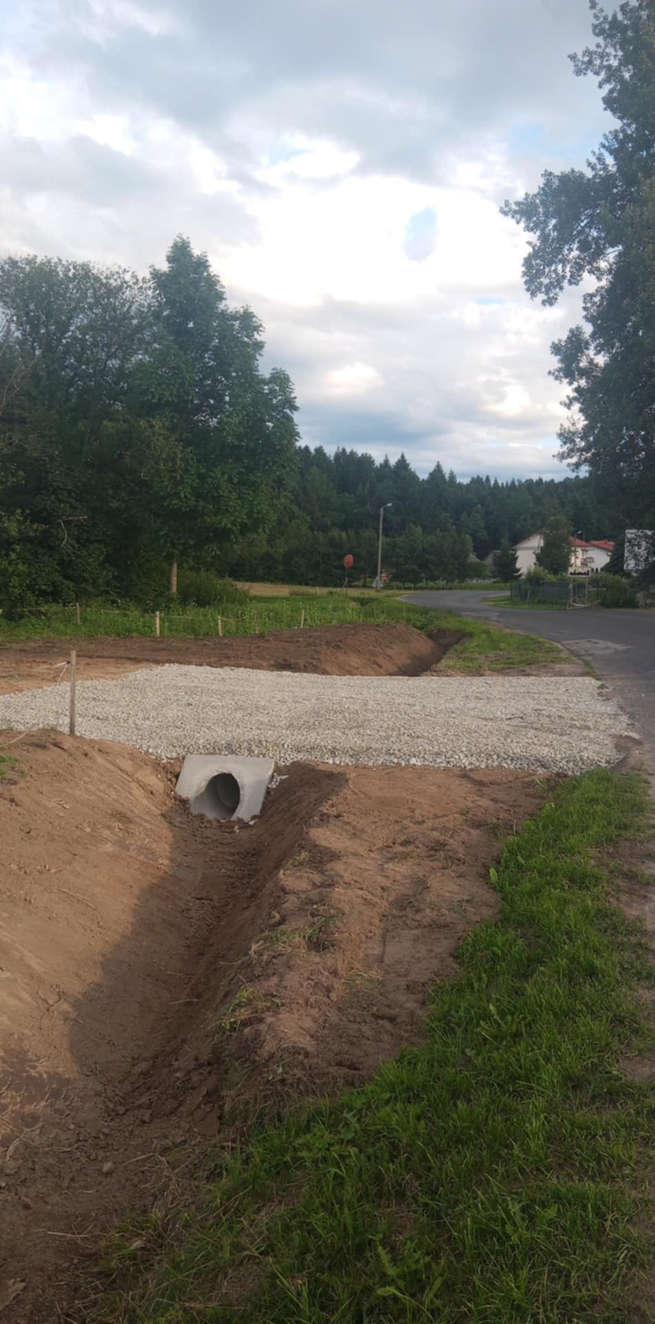 Ukończony drenaż z betonową rurą odpływową, obsypany żwirem, na tle zieleni i pochmurnego nieba. Widoczne ślady prac ziemnych.