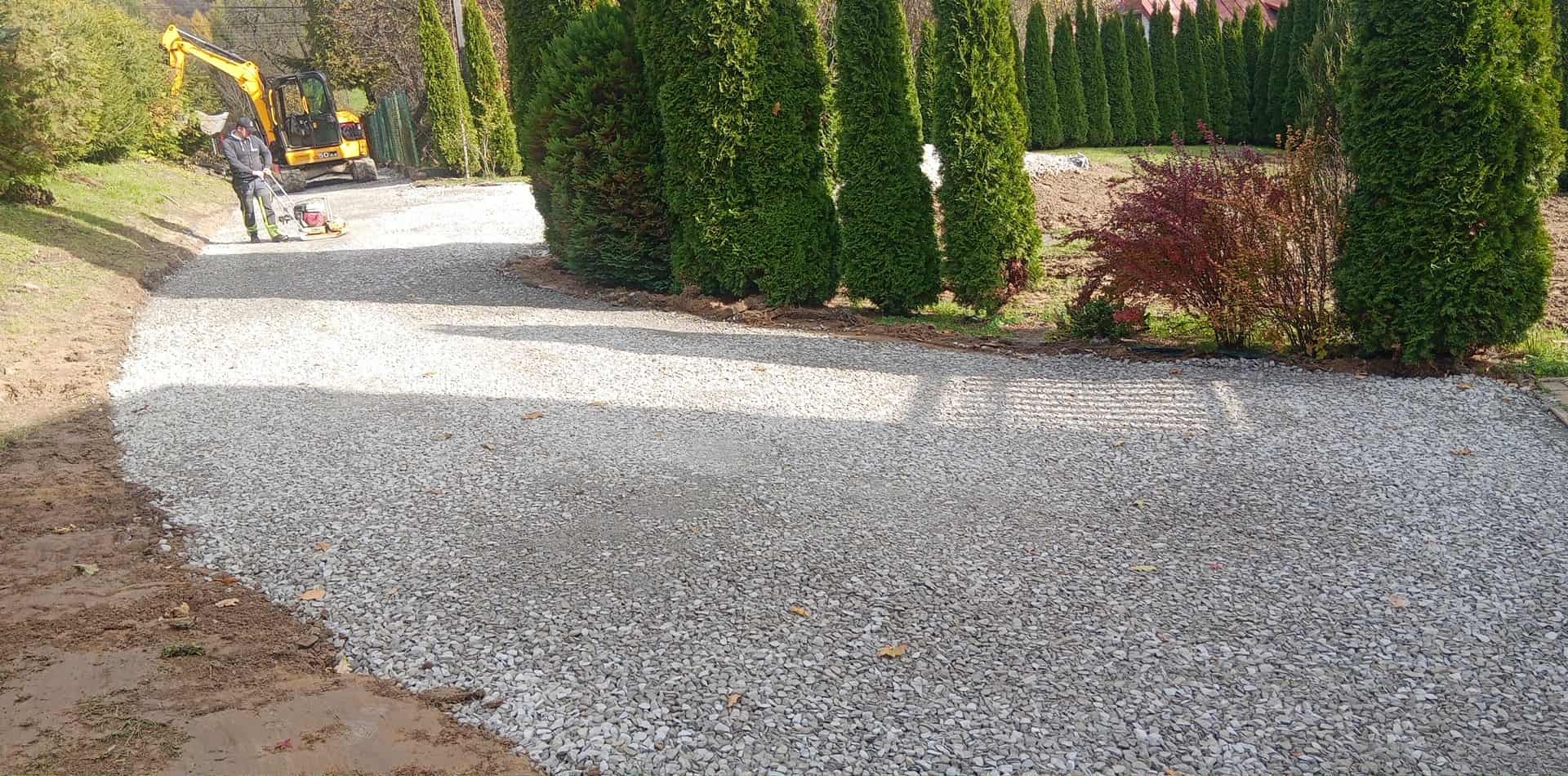 Żwirowa ścieżka w ogrodzie, ubijana przez pracownika z użyciem zagęszczarki. W tle widoczna koparka i rząd tuj.