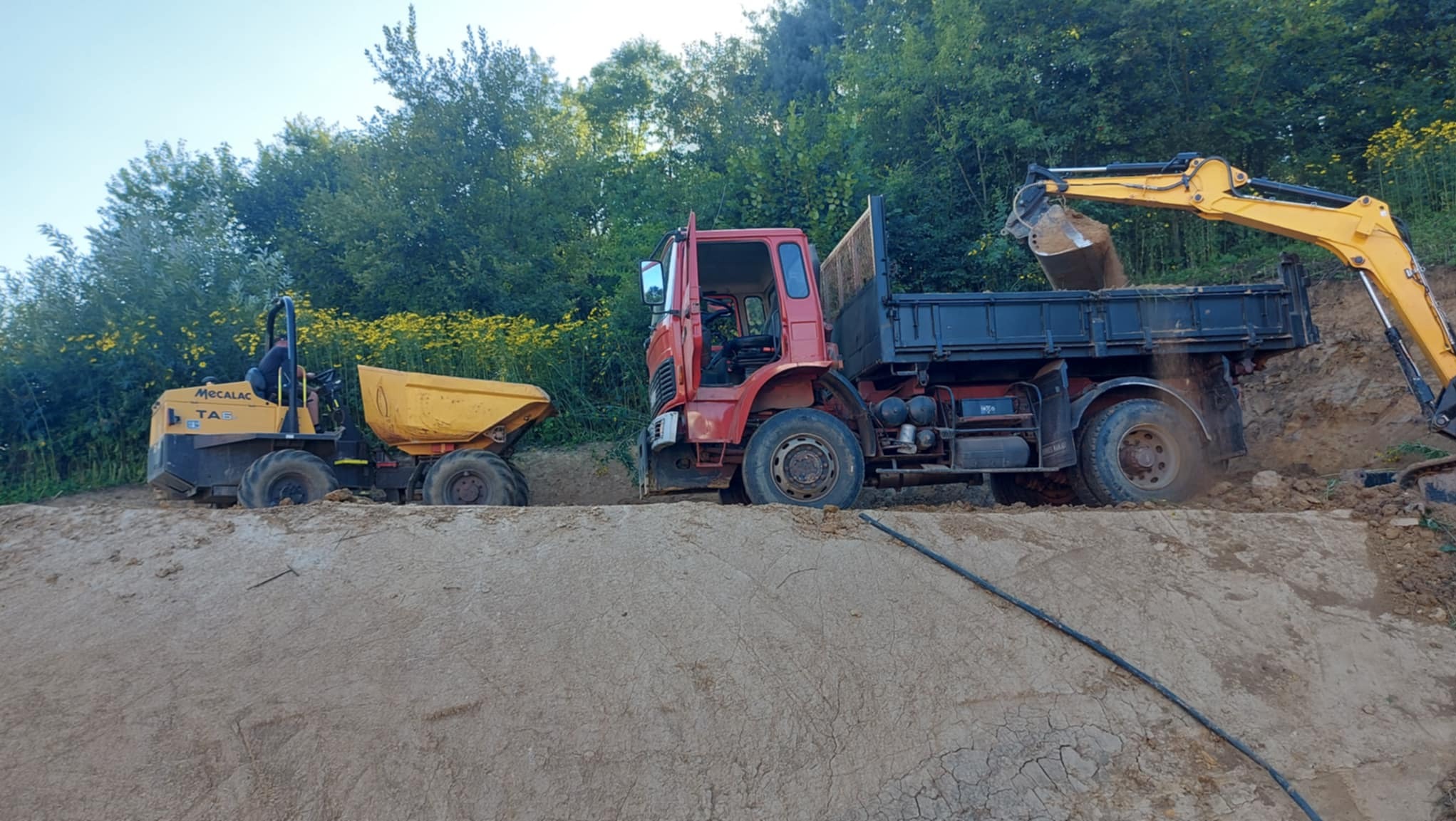 Żółta koparka nabiera ziemię i wsypuje ją do czerwonej ciężarówki na tle zieleni. Obok stoi mała wywrotka. Prace ziemne w toku.