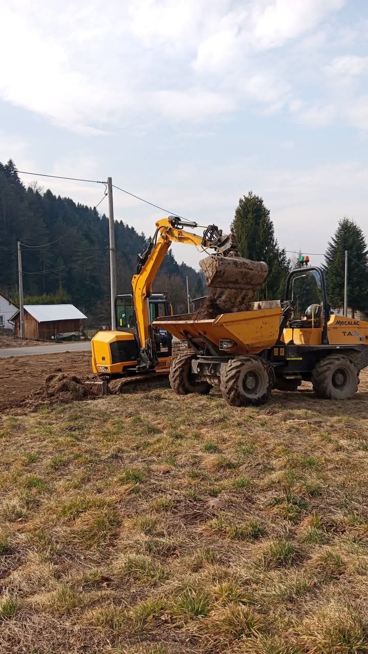 Żółta koparka ładuje ziemię do wywrotki na placu budowy. W tle widoczny las i budynek. Prace ziemne w toku, sprzęt budowlany w akcji.