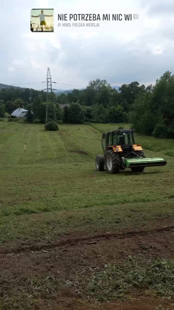 Traktor z kosiarką rotacyjną na polu po koszeniu trawy, widoczne ślady opon i ścięta trawa. W tle słup wysokiego napięcia i zieleń drzew.