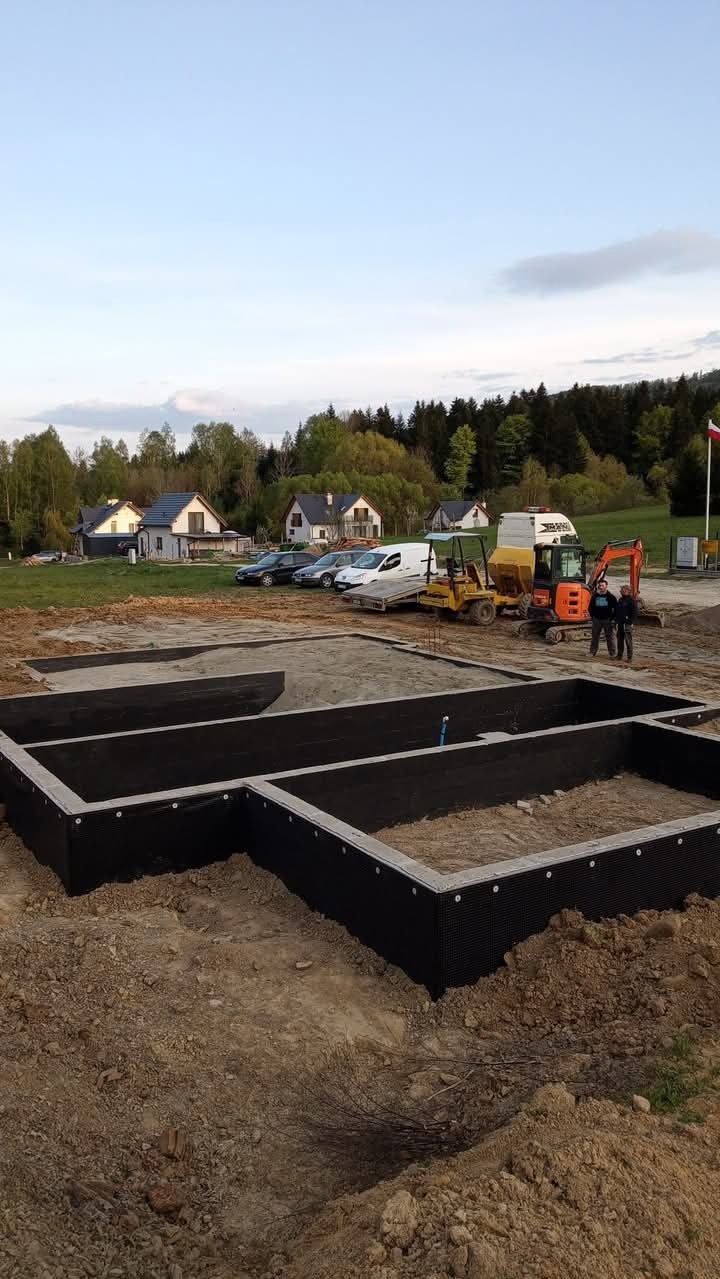 Zabezpieczony fundament domu jednorodzinnego z widoczną hydroizolacją, na tle krajobrazu z domami i sprzętem budowlanym. Dwóch mężczyzn stoi obok pomarańczowej mini koparki.