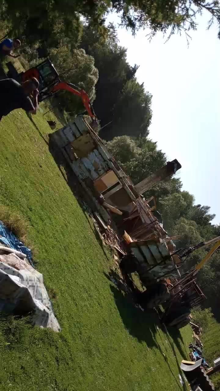 Demolka budynku mieszkalnego przez koparkę na zielonym terenie. Widoczne fragmenty konstrukcji i maszyny budowlanej. Prace rozbiórkowe w toku.