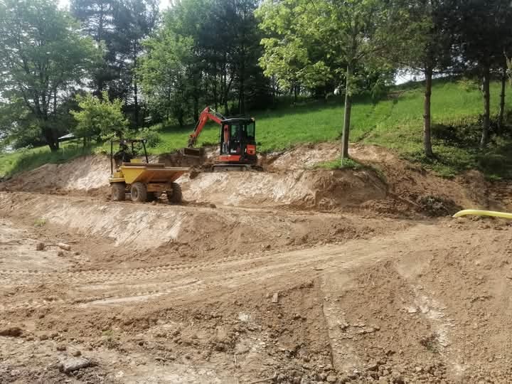 Mini koparka i wywrotka na placu budowy podczas niwelacji terenu. Widoczne ślady opon na ziemi i zielone wzgórze w tle. Prace ziemne w toku.