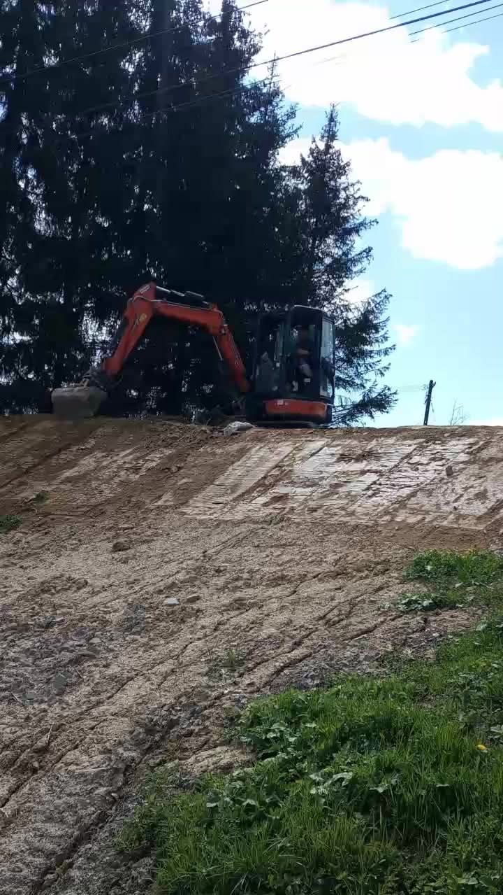 Pomarańczowa mini koparka na nierównym, piaszczystym terenie z zieloną trawą w tle, wykonująca prace ziemne w słoneczny dzień.