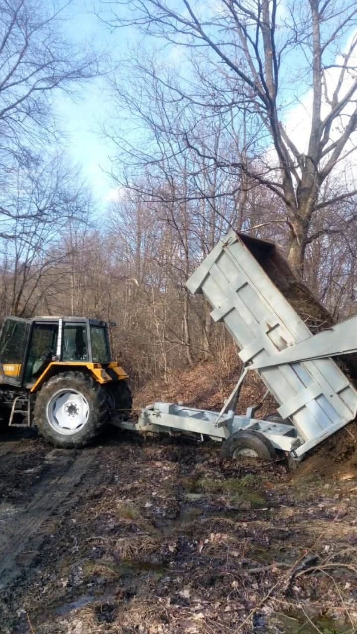 Żółty traktor z przyczepą wywrotką na błotnistym terenie, prawdopodobnie podczas wywozu ziemi. W tle drzewa bez liści i błękitne niebo.