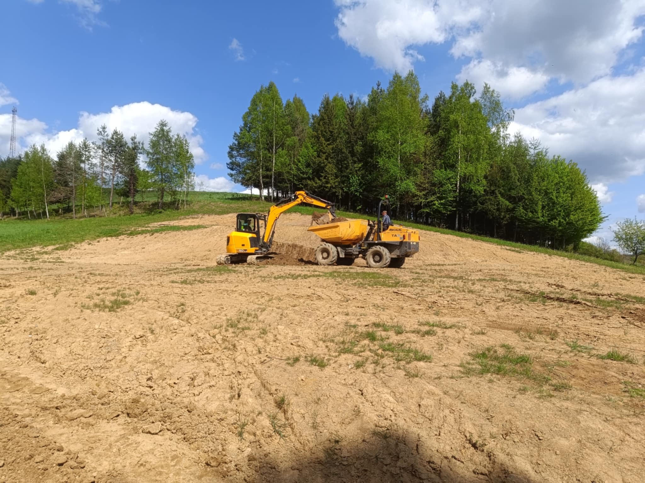 Żółta koparka ładuje ziemię na wywrotkę na wzgórzu, w tle drzewa i błękitne niebo z chmurami. Prace ziemne w toku, widok z daleka.