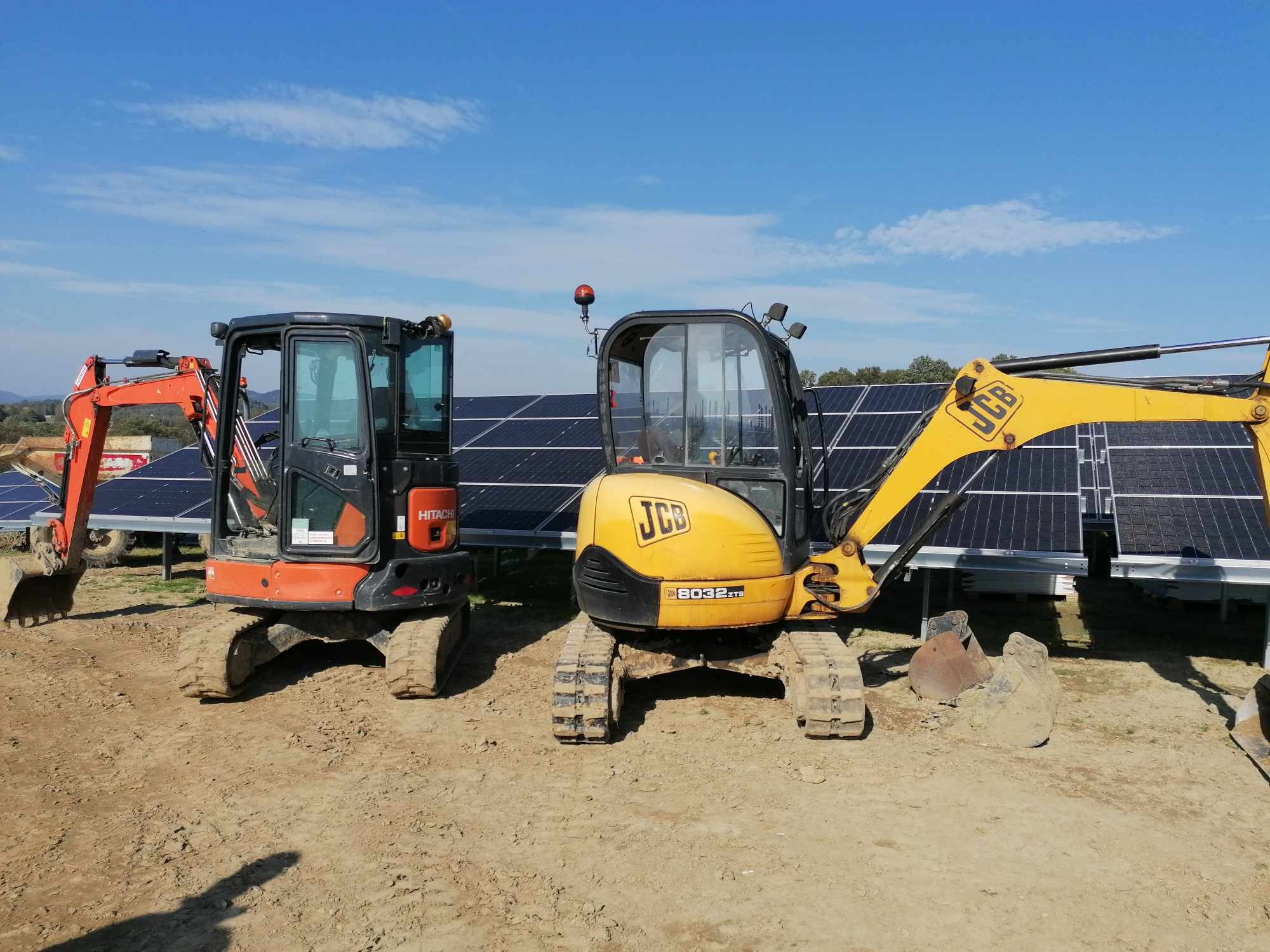 Dwa mini-koparki (pomarańczowa Hitachi i żółta JCB) stoją na ziemi przed rzędem paneli słonecznych pod błękitnym niebem. Widoczne ślady użytkowania.