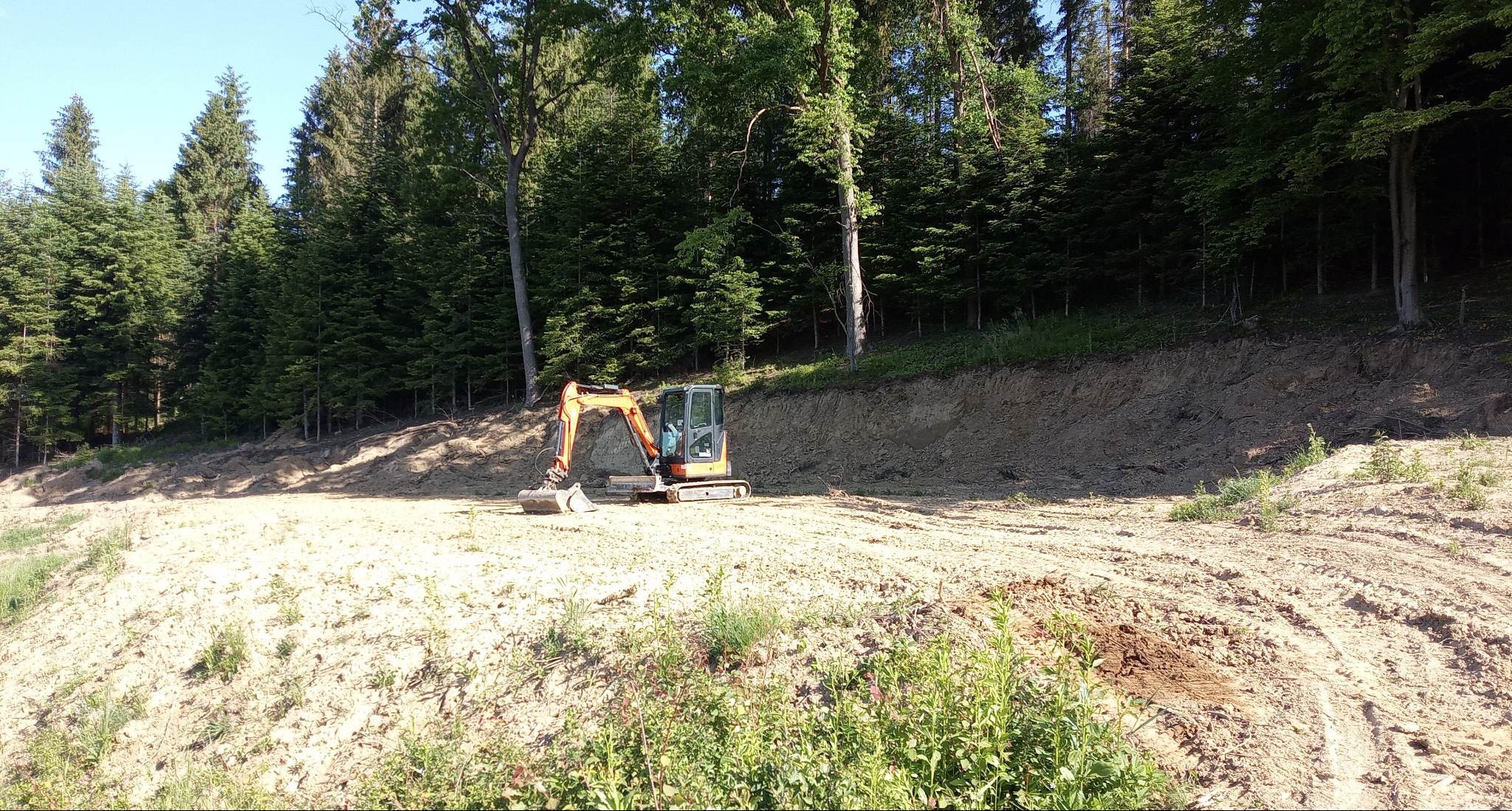 Mała koparka na terenie po niwelacji, z widocznym skarpowaniem i młodym lasem w tle. Ślady opon na piasku.