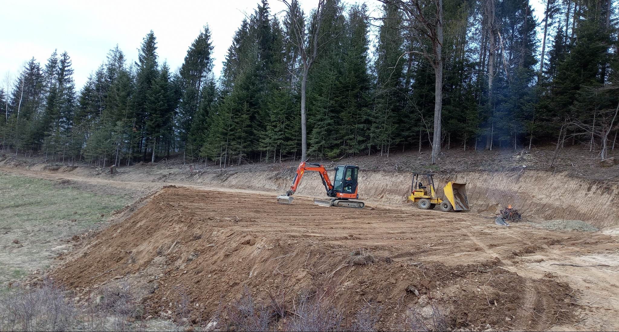 Pomarańczowa mini koparka i żółta ładowarka na wyrównanym terenie z widocznymi śladami pracy, w tle las iglasty i niewielkie ognisko.