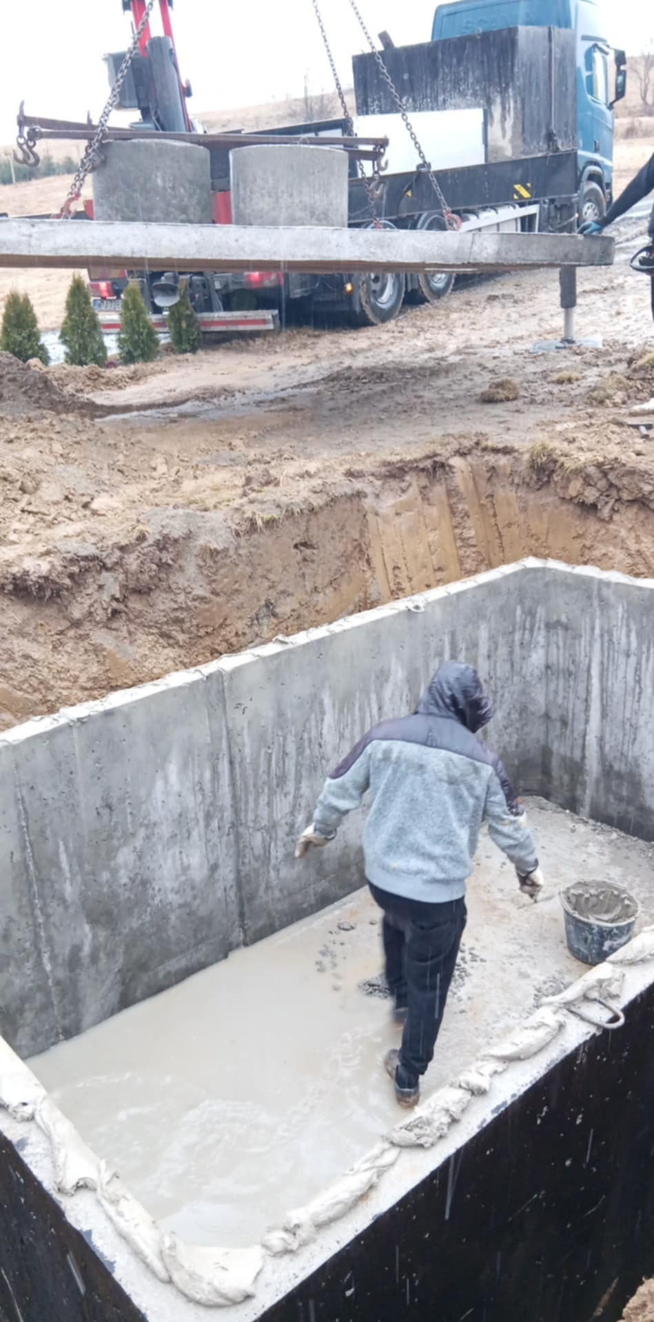 Betonowy zbiornik retencyjny w wykopie, z pracownikiem w środku. Na górze dźwig z betonowymi kręgami. W tle ciężarówka i drzewa.