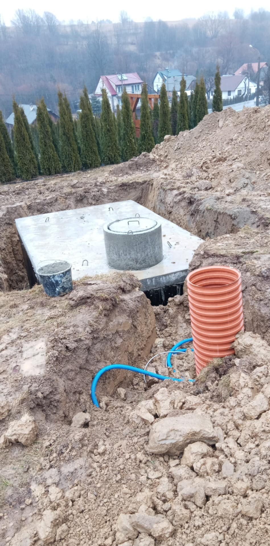 Betonowa studzienka w wykopie, widoczne rury i kabel. W tle rząd tuj i domy. Prace ziemne w trakcie realizacji.