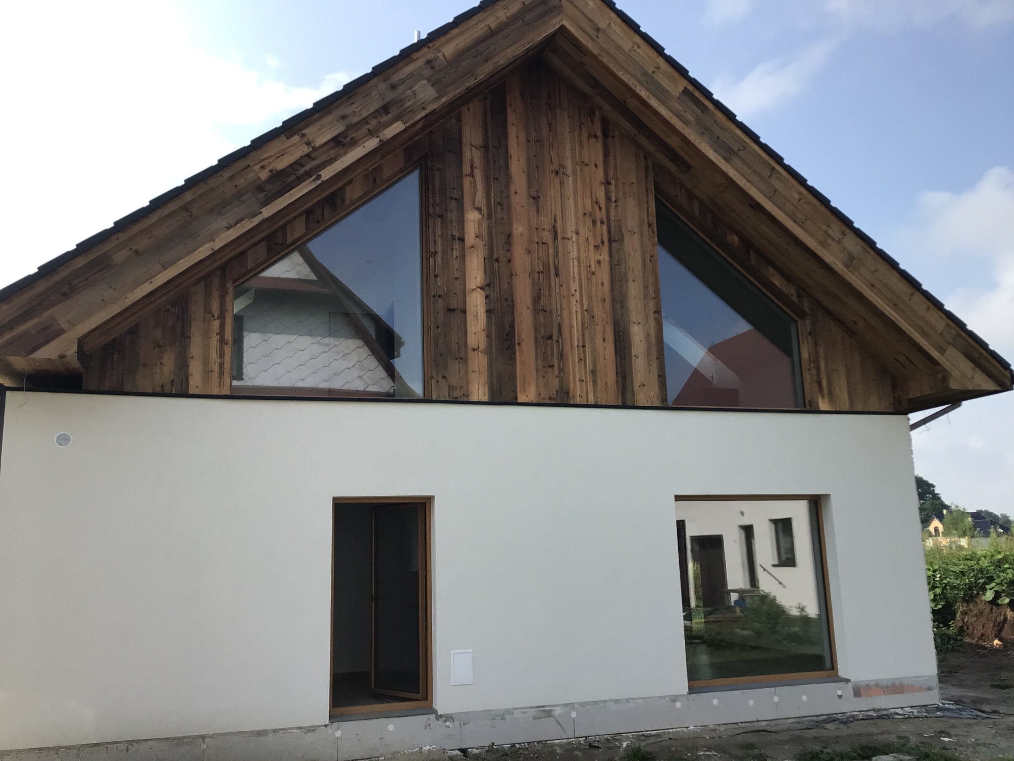 Nowoczesny dom z drewnianą fasadą szczytową i dużymi oknami trójkątnymi. Biała elewacja dolnej części budynku. Widoczne detale wykończenia i surowe otoczenie.