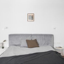 Remton - Minimalistyczna sypialnia: szare łóżko z zagłówkiem, drewniana podłoga, białe ściany z lampkami i szafa. Nad łóżkiem obrazek w ramce. Stonowana kolorystyka.