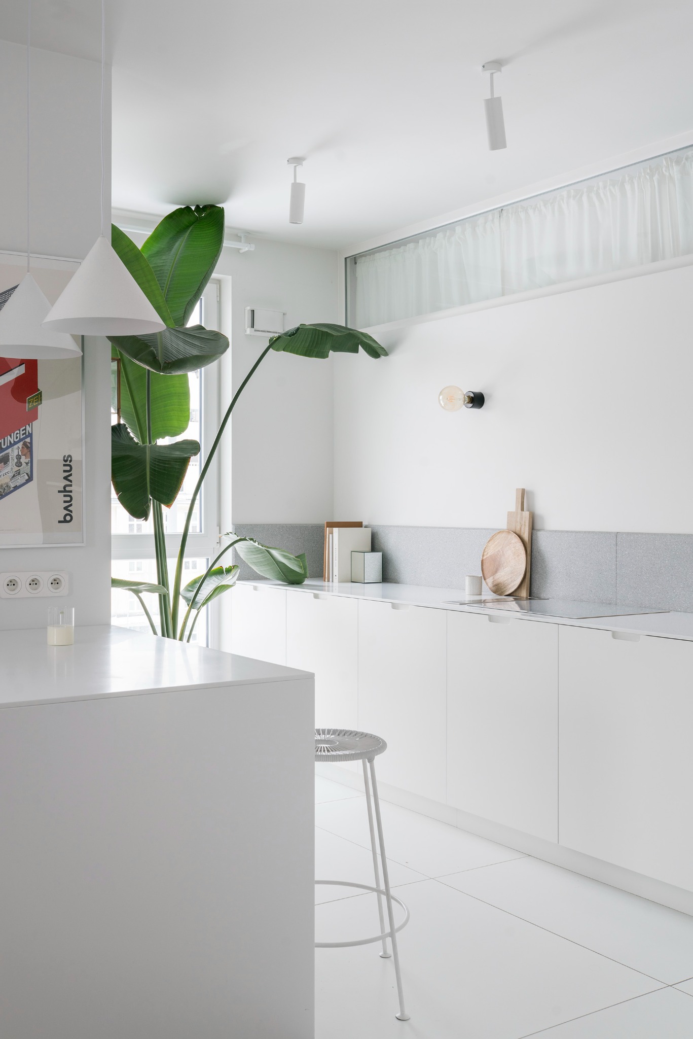 Minimalistyczna, biała kuchnia po remoncie z rośliną strelicją, drewnianymi deskami i plakatem Bauhaus. Widoczne gniazdka elektryczne i krzesło barowe.