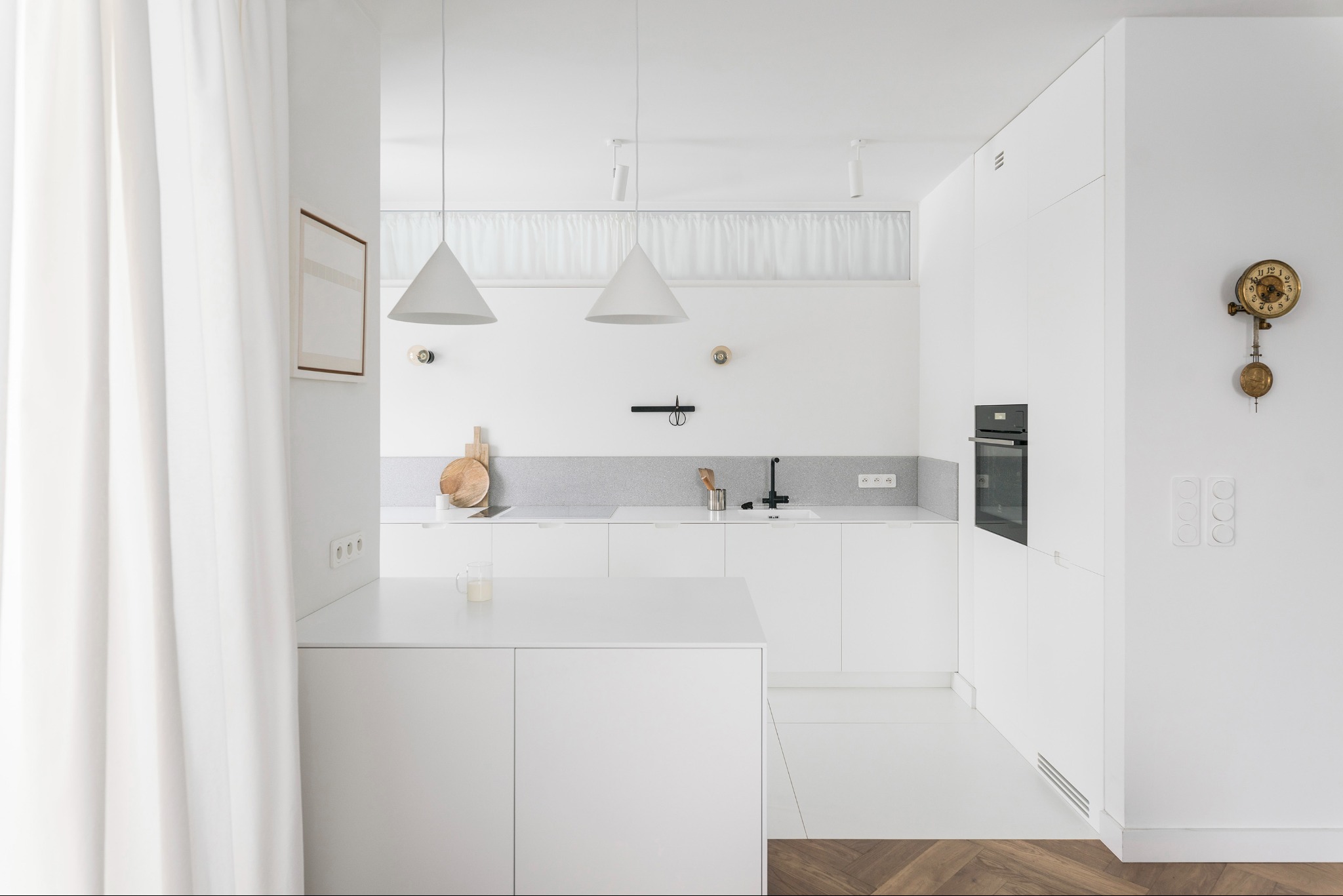 Minimalistyczna, biała kuchnia z drewnianą podłogą w jodełkę, oświetlona wiszącymi lampami. Na ścianie dekoracyjny zegar.