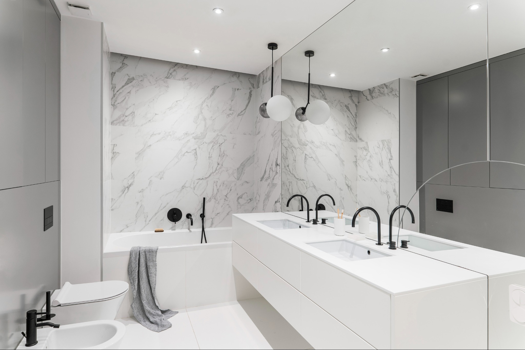 Nowoczesna łazienka z marmurową ścianą, białymi szafkami pod umywalki i czarną armaturą. Minimalistyczny design z wiszącymi lampami kulami i dużym lustrem.