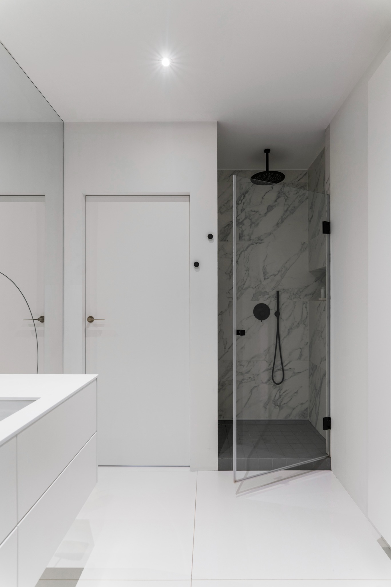 Minimalistyczna łazienka w bieli: kabina prysznicowa z marmurowym wykończeniem, czarna armatura, białe płytki podłogowe i szafka umywalkowa.