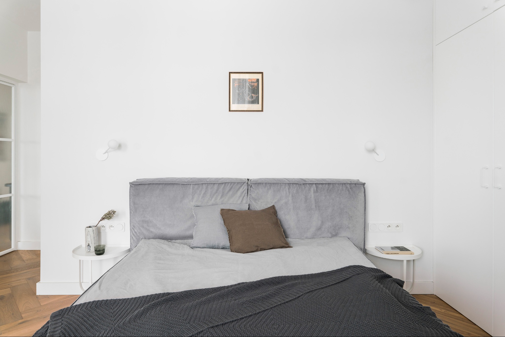 Minimalistyczna sypialnia: szare łóżko z zagłówkiem, drewniana podłoga, białe ściany z lampkami i szafa. Nad łóżkiem obrazek w ramce. Stonowana kolorystyka.