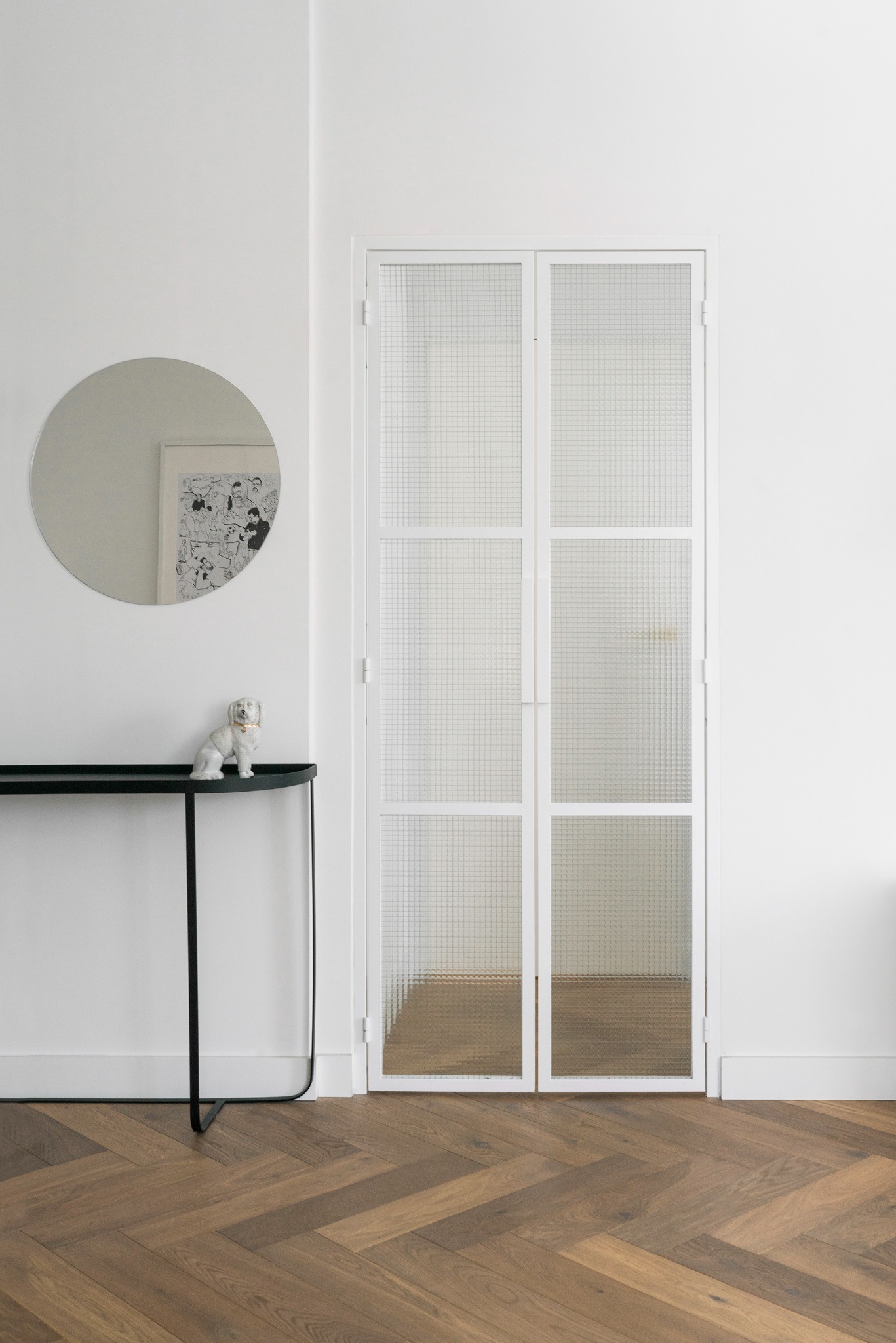Nowoczesne wnętrze: parkiet w jodełkę, białe ściany, przeszklone drzwi z białymi ramami, okrągłe lustro i czarna konsola z figurką psa.