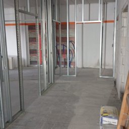 Katarzyna Lorenc Usługi Budowlane - Metalowa konstrukcja ścian działowych w surowym wnętrzu. Widoczne instalacje, podłoga z płytek i worki z zaprawą. Etap prac wykończeniowych w pomieszczeniu.