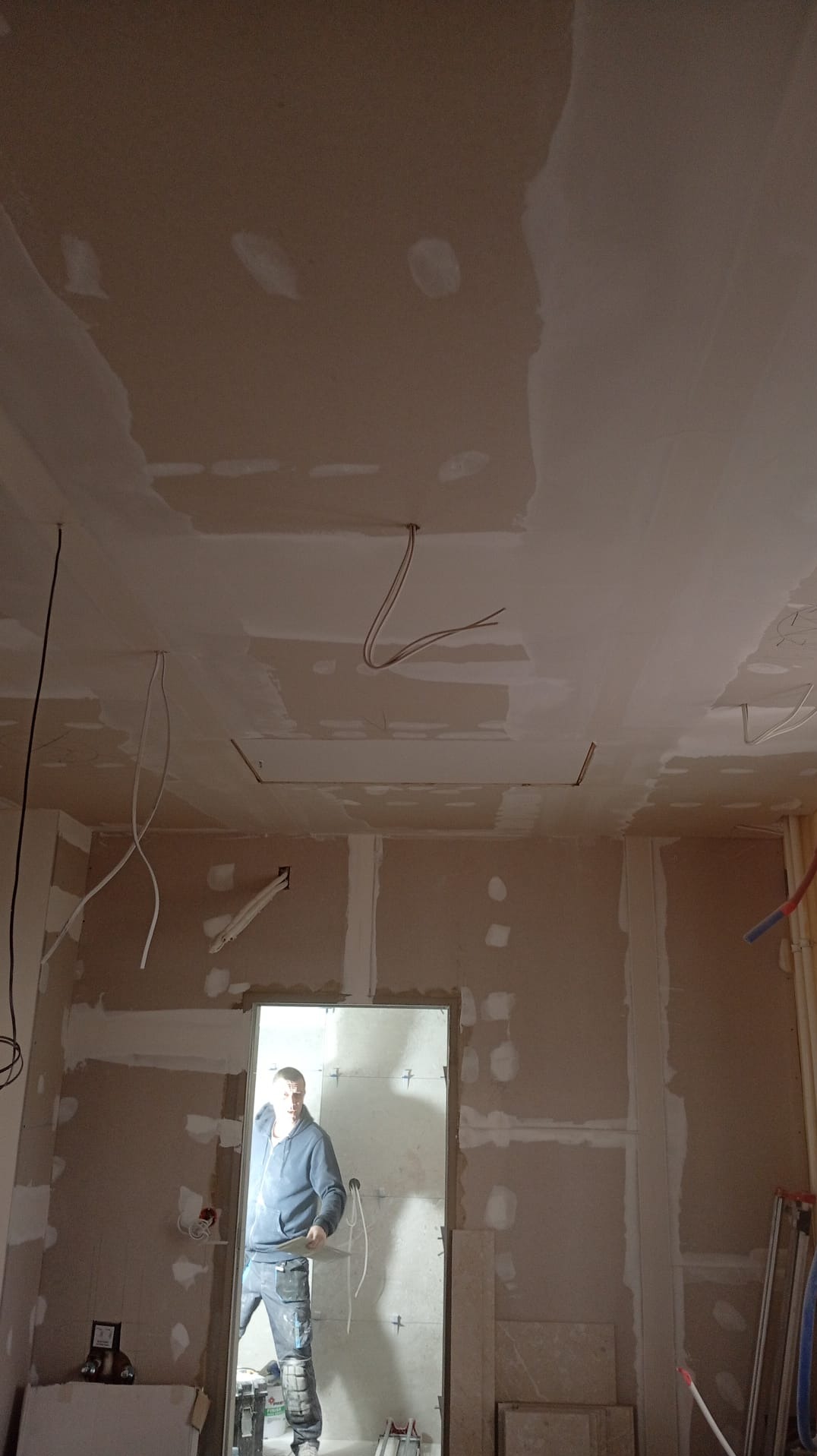 Wnętrze w trakcie prac wykończeniowych: ściany z płyt gipsowo-kartonowych, widoczne przewody elektryczne, pracownik w tle. Ujęcie z dołu.