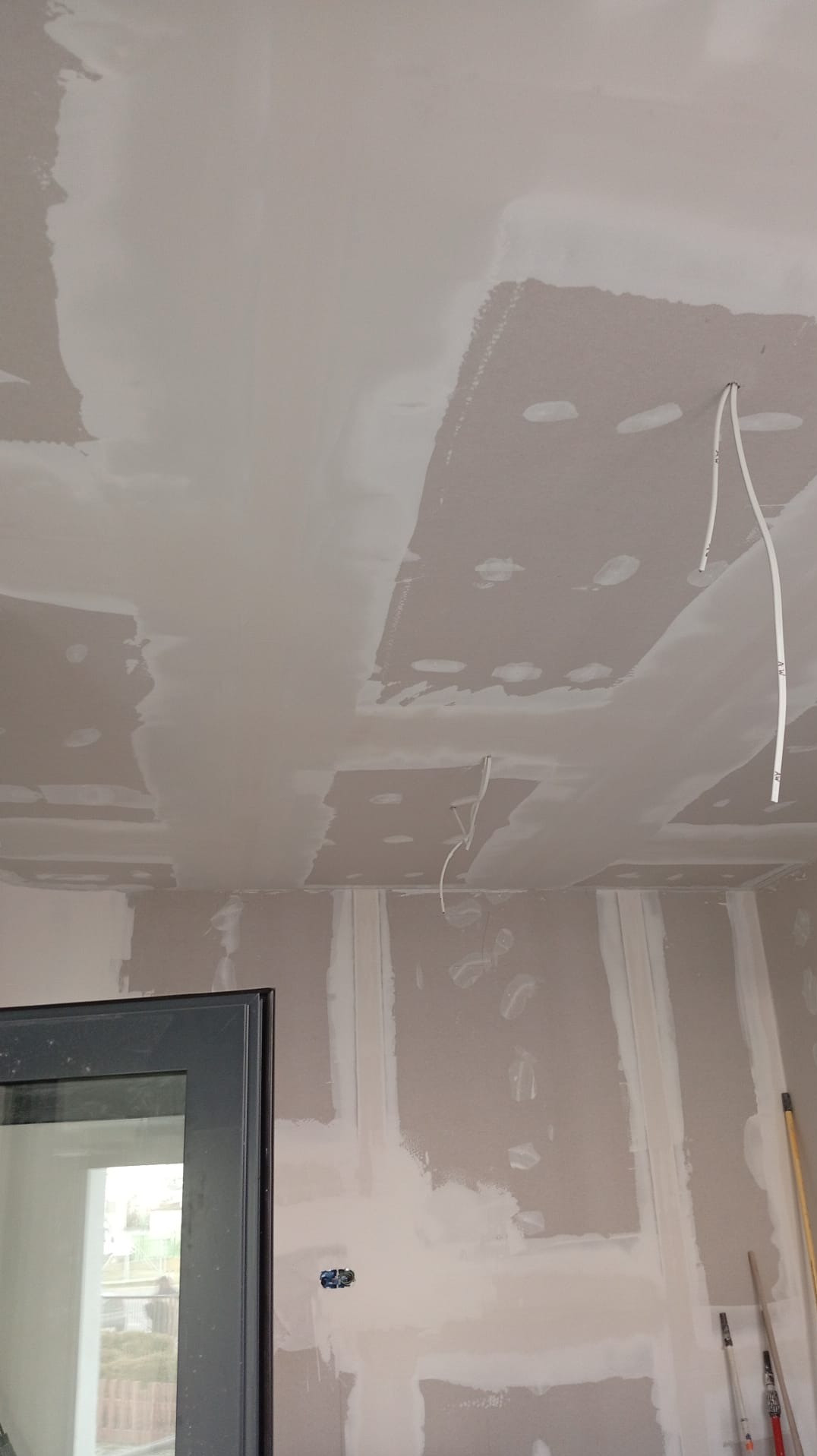 Ściana i sufit po szpachlowaniu, widoczne kable elektryczne. Prace wykończeniowe w pomieszczeniu z oknem. Ślady po łatach na suficie.