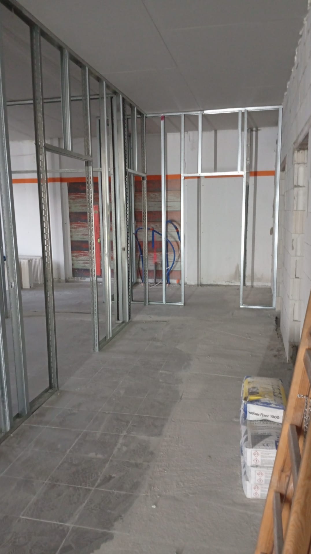 Metalowa konstrukcja ścian działowych w surowym wnętrzu. Widoczne instalacje, podłoga z płytek i worki z zaprawą. Etap prac wykończeniowych w pomieszczeniu.