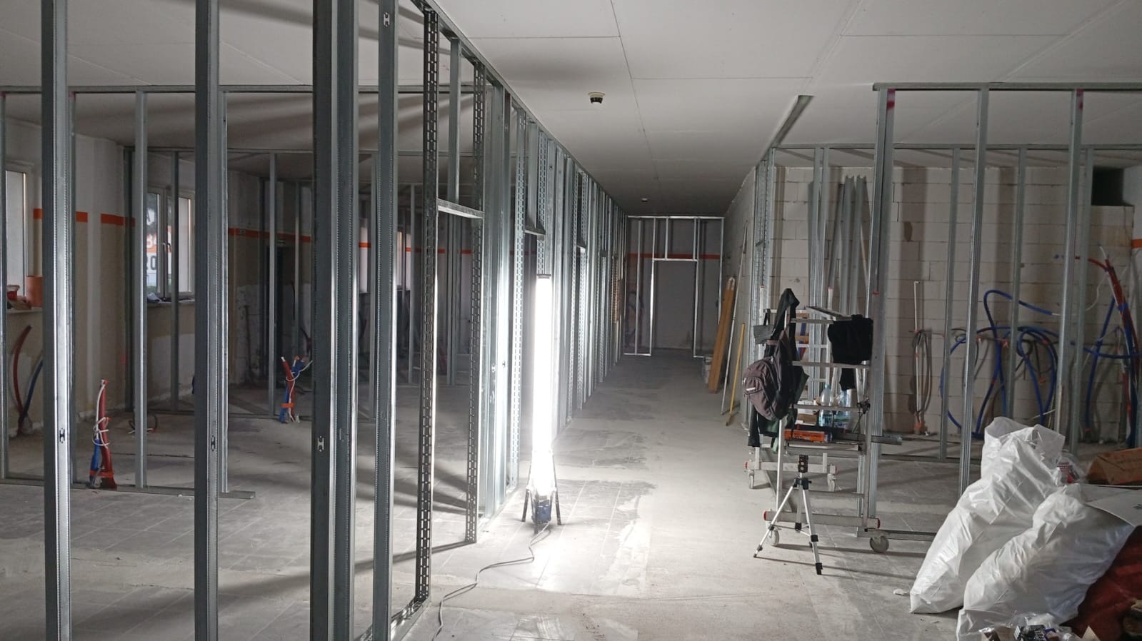 Korytarz w trakcie montażu ścian działowych z płyt gipsowo-kartonowych na metalowym stelażu. Widoczne instalacje, narzędzia i oświetlenie budowlane.