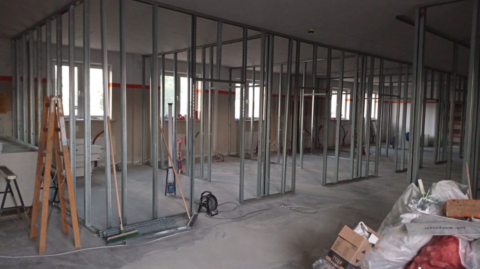 Montaż metalowej konstrukcji ścian działowych w surowym wnętrzu. Widoczne profile, drabina, narzędzia i materiały budowlane. Etap prac wykończeniowych w pomieszczeniu z oknami.