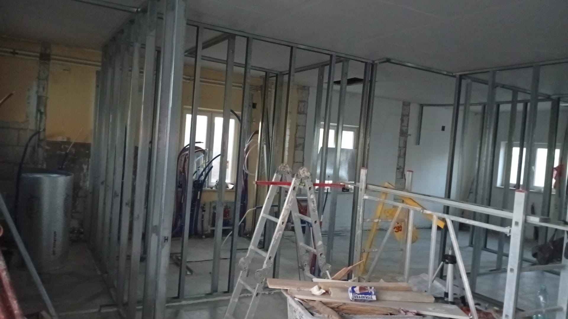 Metalowa konstrukcja ścian działowych w trakcie montażu w remontowanym pomieszczeniu. Widoczne okna, drabina i materiały budowlane. Etap prac przed płytami G-K.