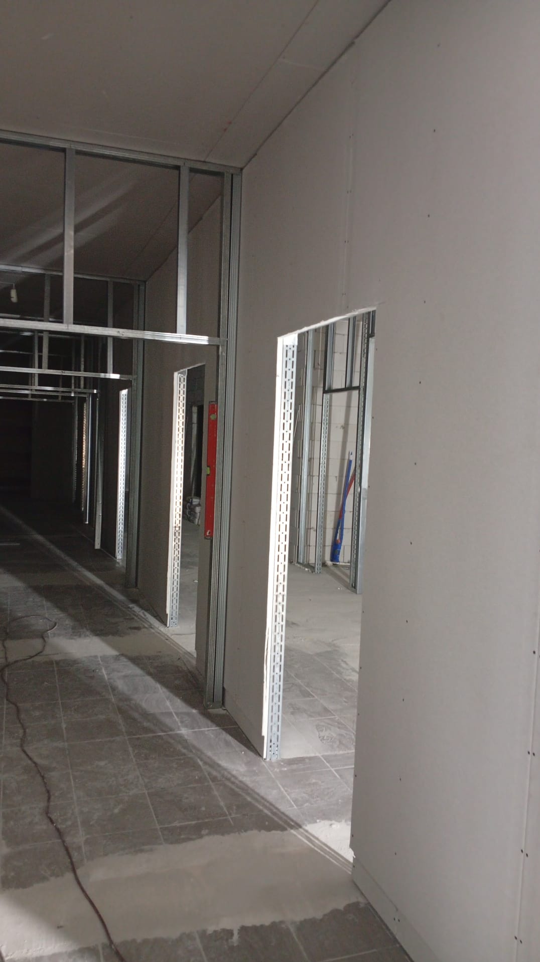 Korytarz w trakcie montażu ścian działowych z płyt karton-gips na metalowym stelażu. Widoczny poziomica i fragment posadzki z płytek. Surowy stan wykończenia.