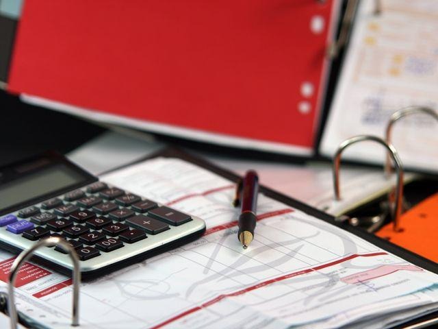 Kalkulator, długopis i otwarty segregator z dokumentami na biurku. Widoczne czerwone segregatory w tle. Ujęcie z góry, oświetlenie biurowe.