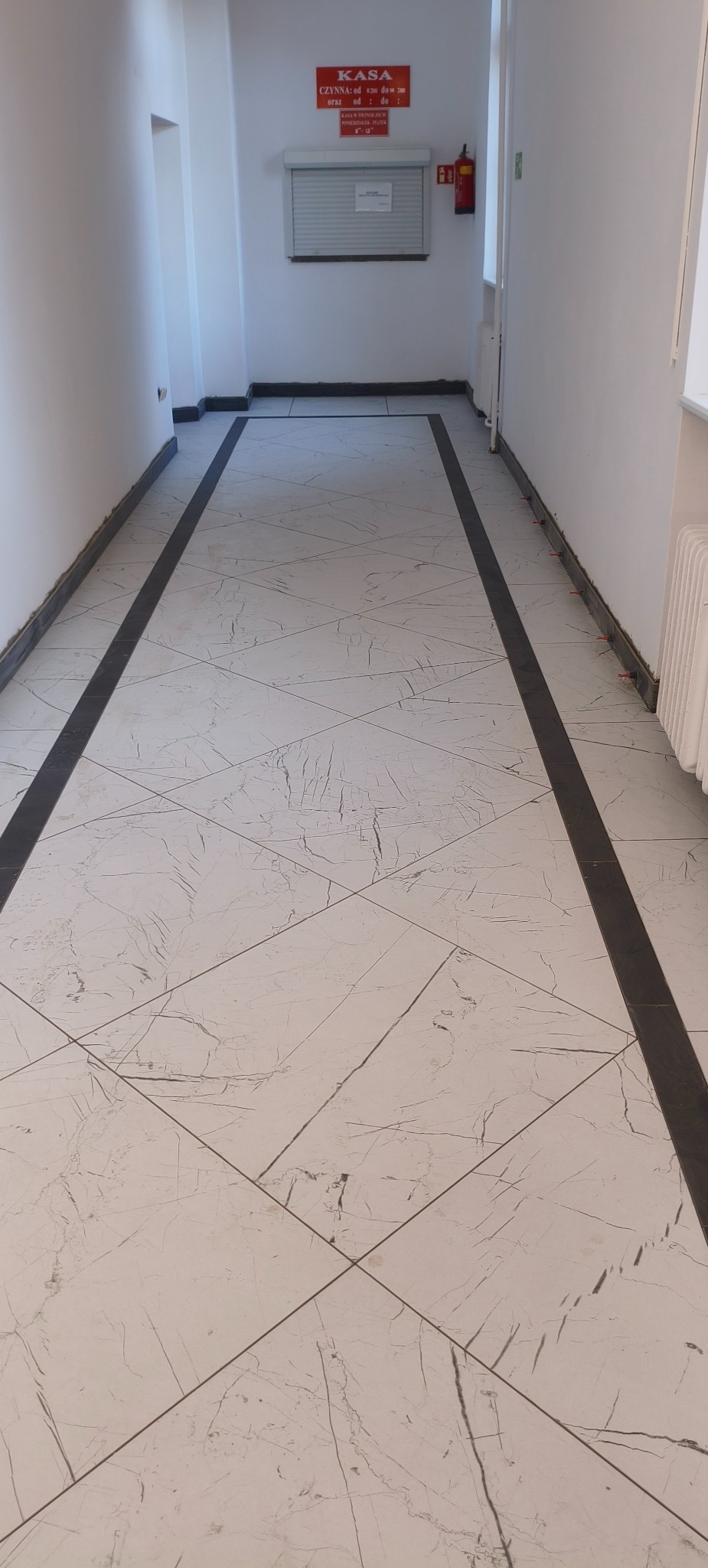 Białe płytki podłogowe z szarymi żyłkami, ułożone w romby, obramowane czarnymi pasami wzdłuż ścian w korytarzu. Widoczny fragment grzejnika i tabliczka informacyjna.