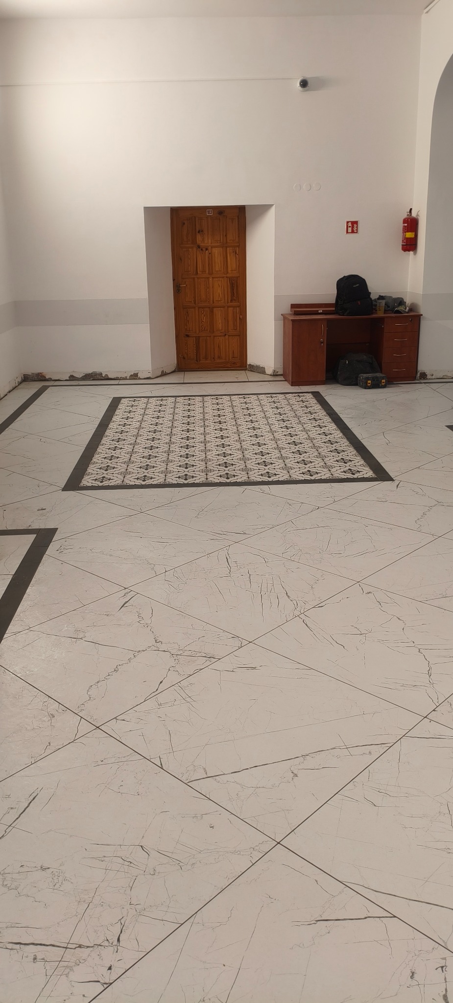 Wykończona podłoga z płytek imitujących marmur, z dekoracyjnym wzorem w centralnej części. Widoczna drewniana stolarka drzwiowa i biurko z wyposażeniem w tle.