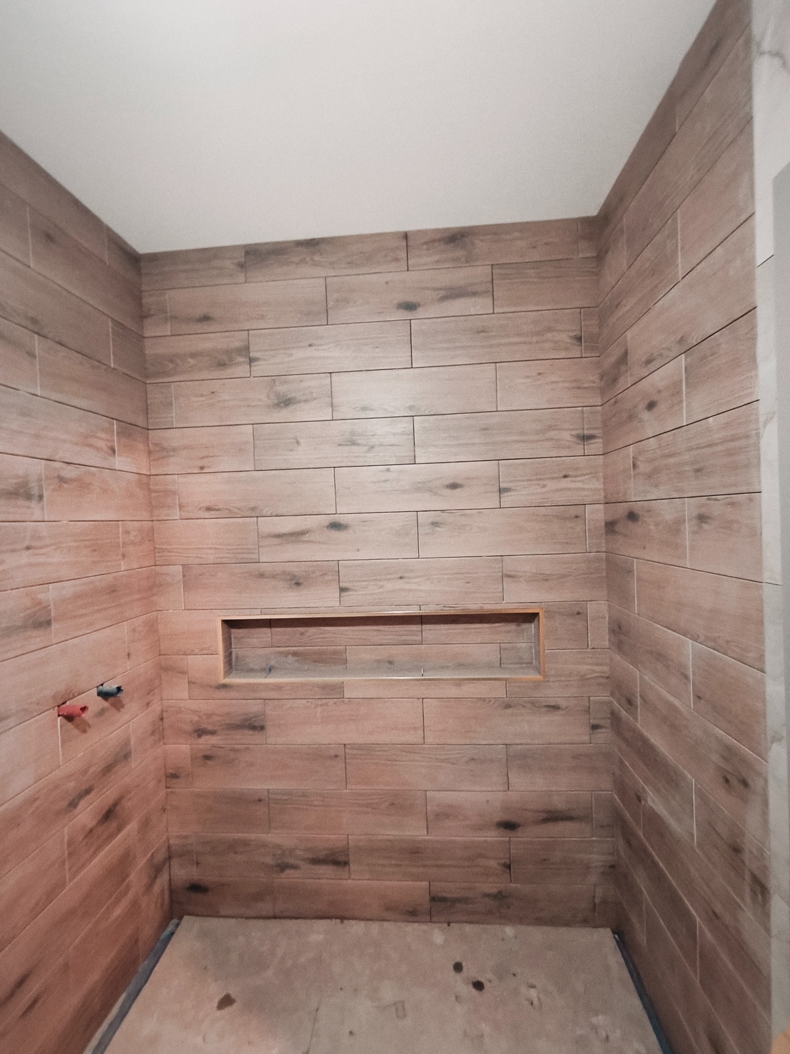 Wykończona kabina prysznicowa z drewnopodobnymi płytkami i wnęką. Widoczne fragmenty instalacji wodnej. Podłoga w trakcie prac wykończeniowych.
